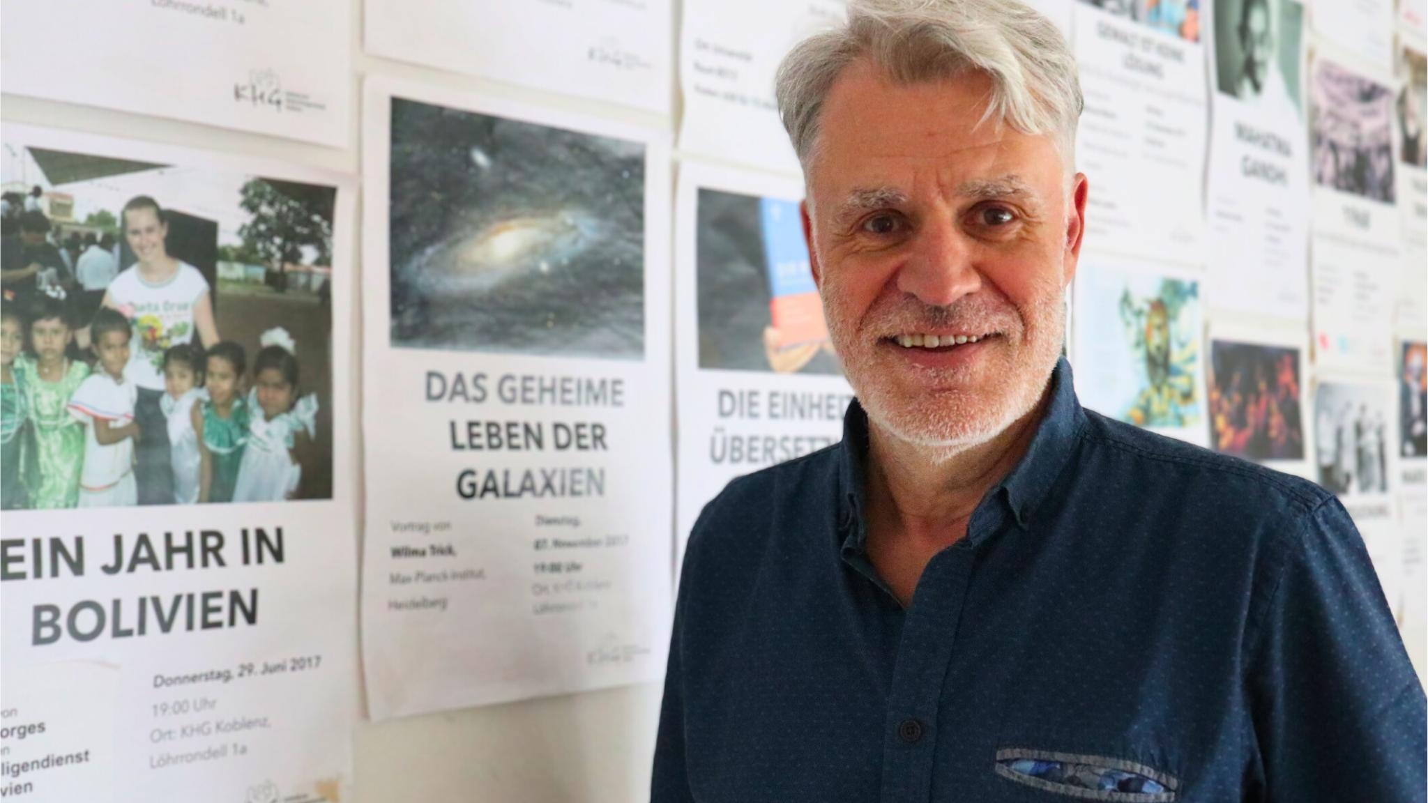 Auf dem Foto steht Guido Groß vor einer Pinnwand mit Veranstaltungsankündigungen der Katholischen Hochschulgemeinde Koblenz. Zu sehen sind sein Oberkörper und der Kopf. Er trägt ein kurzärmliges blaues Hemd, hat graue Haare und einen grauen Drei-Tage-Bart und schaut lächelnd in die Kamera.