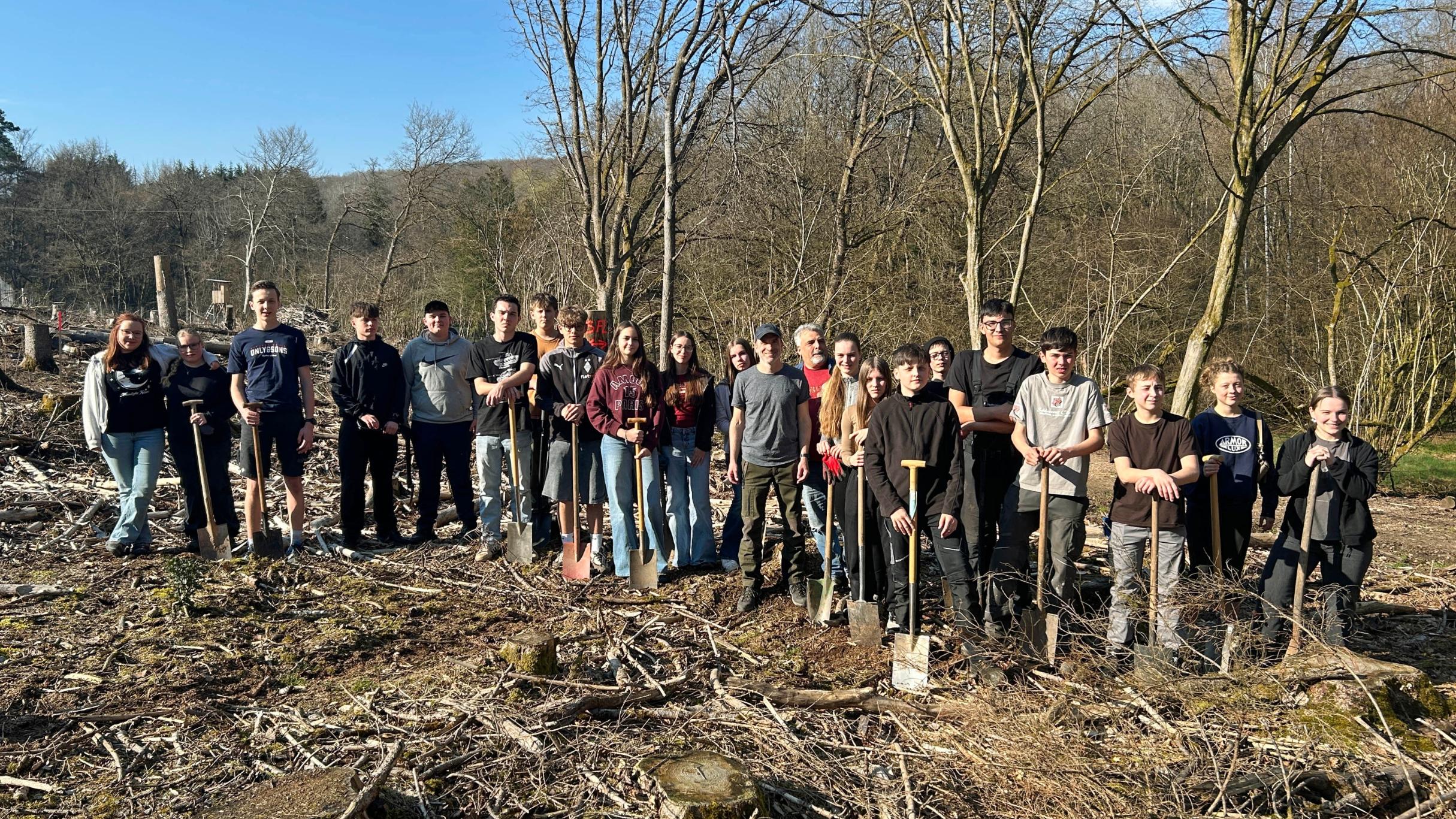 Eine große Gruppe Jugendlicher und Erwachsener steht auf einer offenen, kahlen Waldfläche zwischen dünnen, blattlosen Bäumen. Der Boden ist mit Ästen, Rindenschollen und Holzresten bedeckt. Viele der Teilnehmenden halten Spaten in den Händen und stehen in einer Reihe für ein Gruppenfoto. Im Hintergrund sind mehr Bäume, sanfte Hügel und ein blauer Himmel mit etwas Sonnenschein zu sehen.