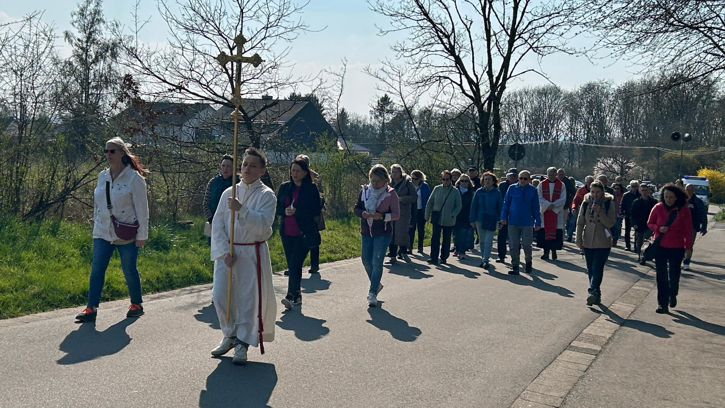 Eine größere Gruppe von Menschen geht bei sonnigem Wetter auf einer asphaltierten Straße zwischen Wiesen und Bäumen. An der Spitze der Prozession trägt eine Person ein großes Vortragekreuz. Hinter dieser Person folgt eine dicht geschlossene Gruppe von Teilnehmenden, einige in Jacken, andere mit Taschen oder Rucksäcken. Rechts und links des Weges stehen noch kahle Bäume und frühe Frühlingssträucher. Im Hintergrund sind einige Häuser und ein freies Feld zu sehen. Die Szene wirkt wie eine gemeinschaftliche religiöse Prozession oder ein gemeinsamer Weg durch die ländliche Umgebung.