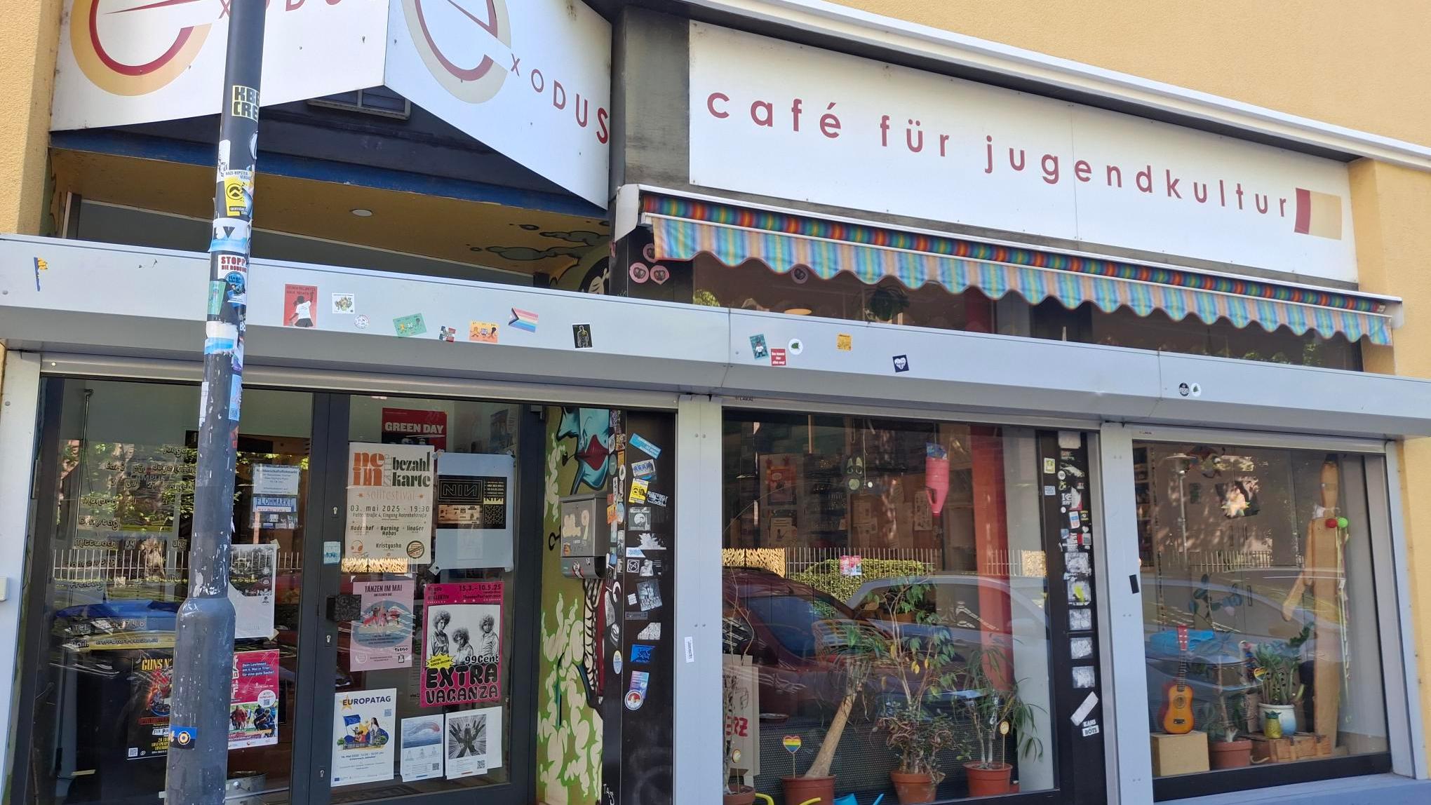Eingang des ‚Café Exodus – Café für Jugendkultur‘ in einem gelben Gebäude. Über dem Eingang hängen zwei Schilder mit dem Schriftzug ‚Exodus‘ und ‚Café für Jugendkultur‘. Vor den großen Fenstern stehen viele Pflanzen, und die Glasflächen sind mit zahlreichen Stickern und Plakaten beklebt. Links befindet sich eine Glastür mit weiteren bunten Plakaten, die auf Veranstaltungen hinweisen. Ein Laternenpfahl steht am linken Bildrand und ist ebenfalls mit Stickern beklebt.