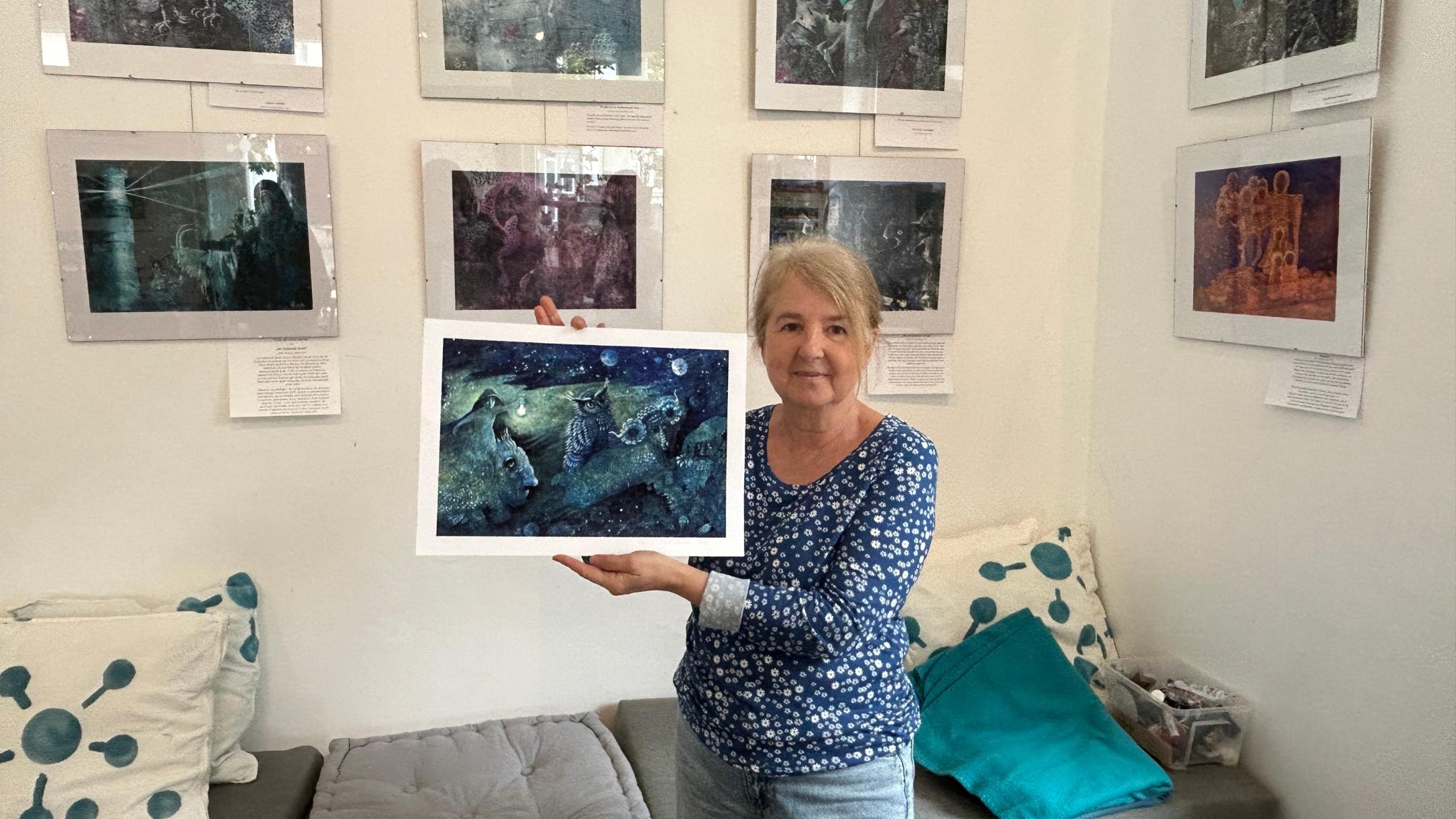 Eine Frau hält eins der von ihr gemalten Bilder in der Hand, weitere hängen im Hintergrund an der Wand.