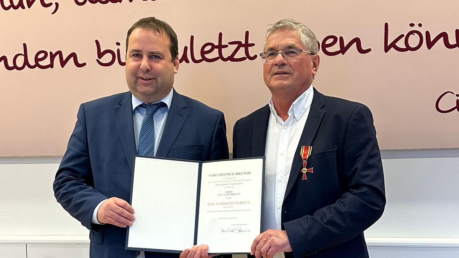 Zwei Personen stehen nebeneinander in einem Innenraum vor einer großen Wand mit dekorativer Schrift in dunkler Farbe. Beide tragen formelle Kleidung. Die Person links (Sozialminister Magnus Jung) hält eine geöffnete Urkunde mit beiden Händen. Die Person rechts (Paul Herrlein) trägt ein Jackett, an dem ein Orden mit rotem Band befestigt ist, und berührt die Urkunde mit einer Hand. Der Boden ist aus hellem Holz, und hinter den beiden verläuft ein weißes Paneel entlang der Wand.